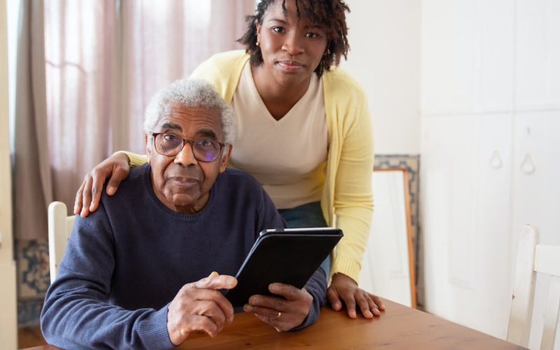 a-woman-standing-beside-the-elderly-man-holding-a-tablet